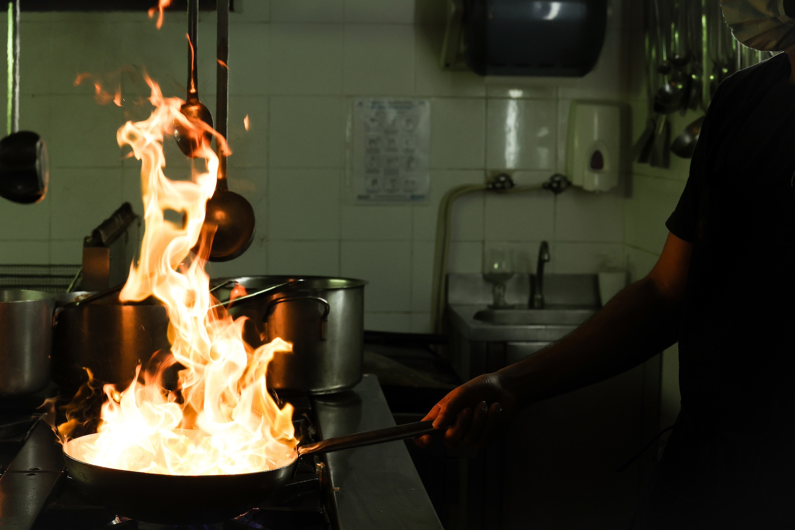 A person holds the handle of a skillet from which flames are emerging, on top of a large industrial range