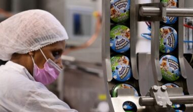 A worker in a hairnet and pink mask operates a production line at a Ben & Jerry's factory, with ice cream pots labelled &quot;Chunky Monkey&quot; visible on the conveyor