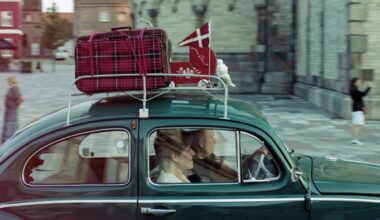 A couple in a green car with red luggage, a small pennant of the Danish flag and a red doll’s pram strapped to the roof, drive through a town