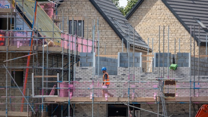 Construction site of new houses in Midsomer Norton, England, with scaffolding and partially built brick walls. A worker wearing a blue hard hat and orange safety vest is visible walking on the scaffolding.