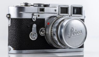 A close-up of a vintage Leica camera with a silver body and black textured grip. The lens cap prominently displays the Leica logo. The camera rests against a white background.