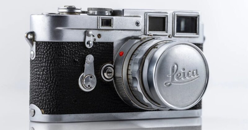 A close-up of a vintage Leica camera with a silver body and black textured grip. The lens cap prominently displays the Leica logo. The camera rests against a white background.
