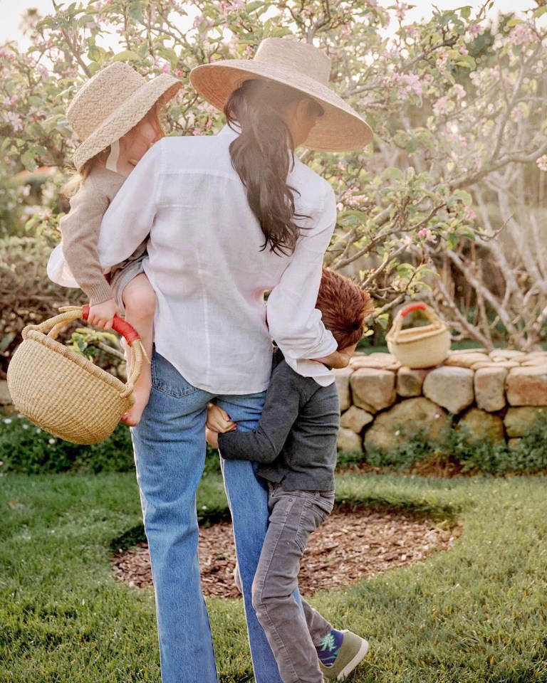 Woman carrying a young child while another child holds onto her leg in a garden.
