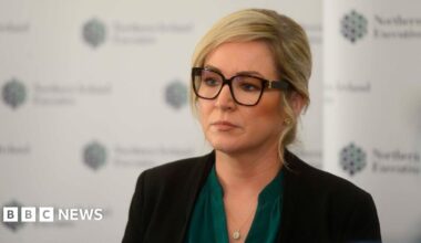 O'Neill did not see McMonagle at Stormont event - report Michelle O'Neill standing in front of a background with the logo of the Northern Ireland Executive. She is wearing glasses, a green top, a silver necklace, and a black blazer. She is looking off camera with a neutral expression.