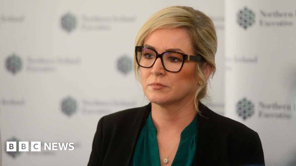O'Neill did not see McMonagle at Stormont event - report Michelle O'Neill standing in front of a background with the logo of the Northern Ireland Executive. She is wearing glasses, a green top, a silver necklace, and a black blazer. She is looking off camera with a neutral expression.