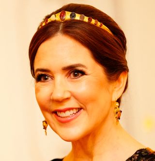 A headshot of Queen Mary wearing a slim gold tiara with stones on it and smiling in front of an ivory backdrop