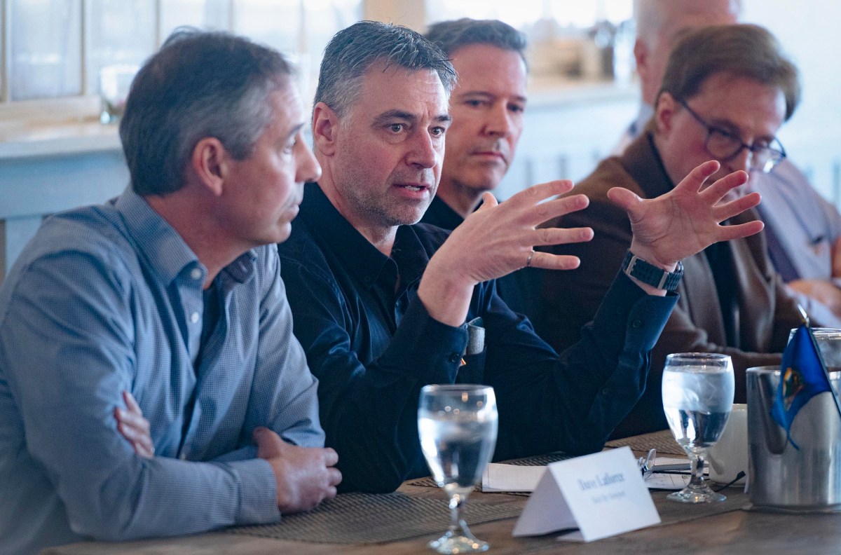A man in a dark shirt speaks animatedly at a table with water glasses and placemats, while others listen attentively.