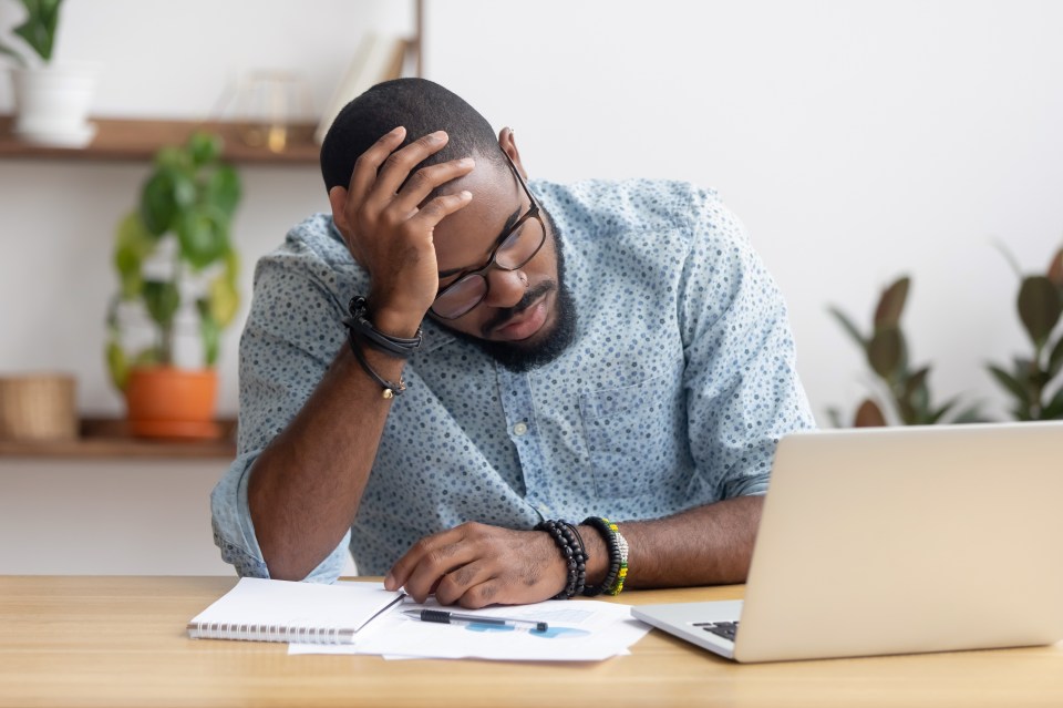 A tired businessman sits at his desk, head in hand, looking at his laptop.