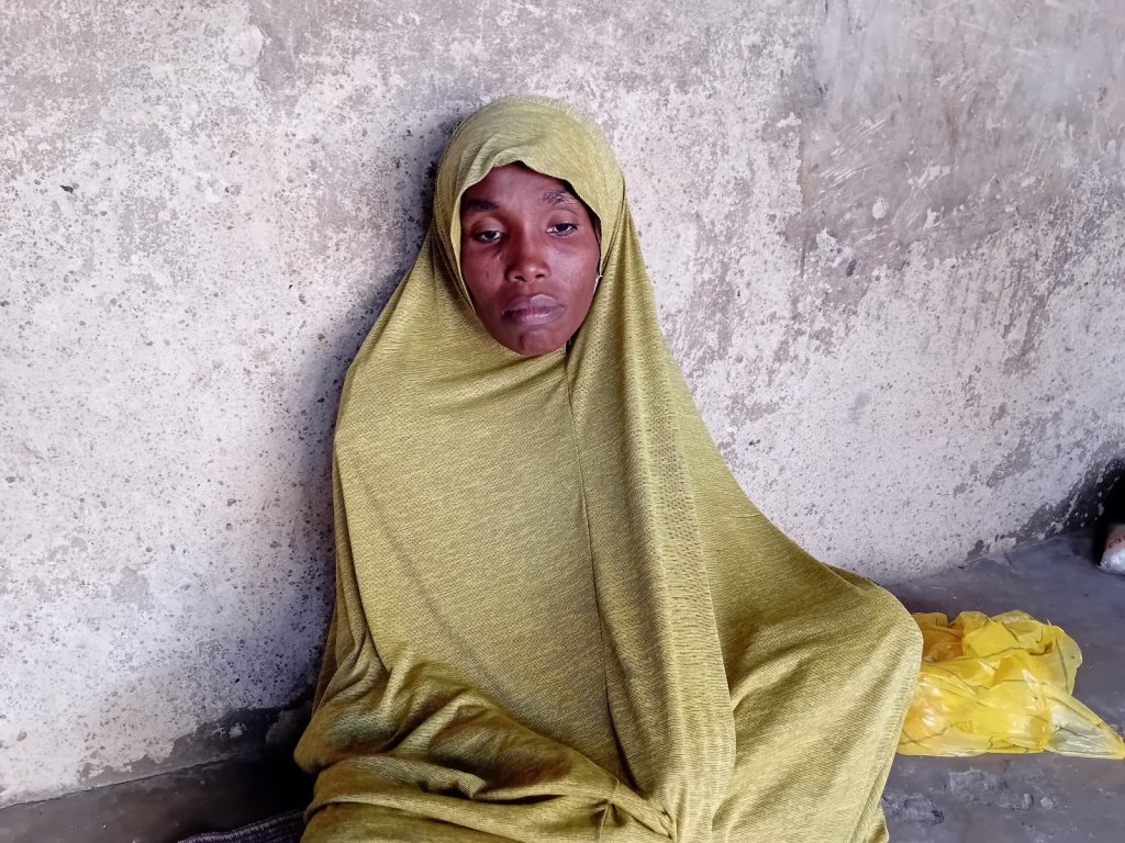 A person in a green hijab sits against a textured gray wall, looking thoughtfully.