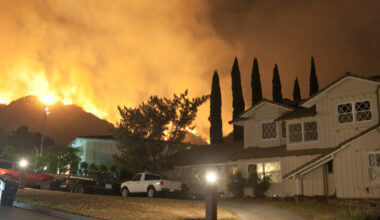 A large wildfire blazes on a hillside behind residential homes at night, illuminating the sky with orange flames and smoke.
