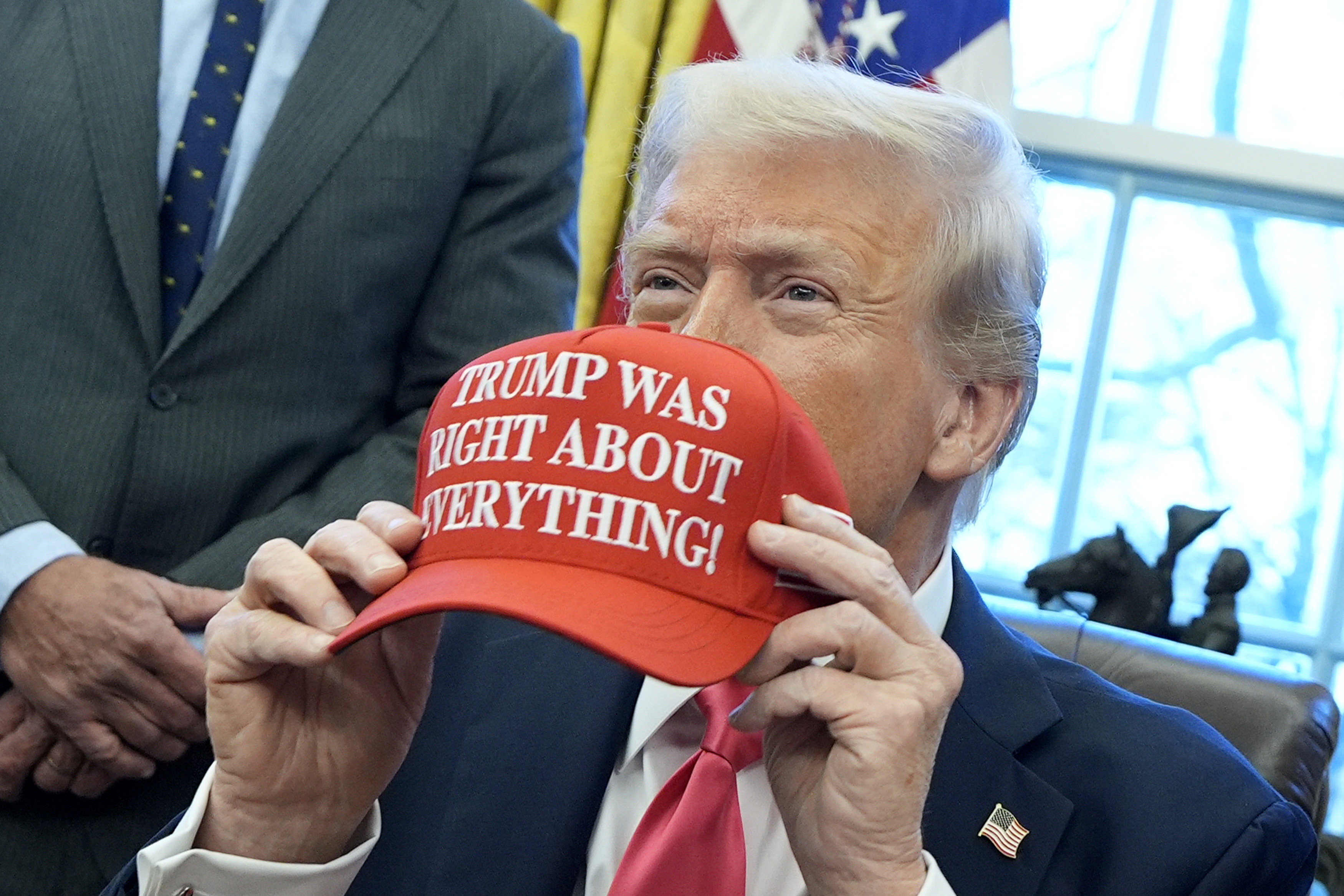 President Trump holding a red hat that reads "Trump Was Right About Everything!" in the Oval Office.