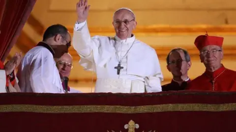 Getty Images Pope Francis shown on the balcony of the Vatican. He is dressed in white and has a large cross around his neck. He is waving to the crowd.