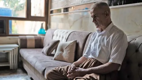 Phil Caller A middle-aged man in white shirts sits contemplatively on a large brown sofa, his hands folded in his lap, as he stares ahead