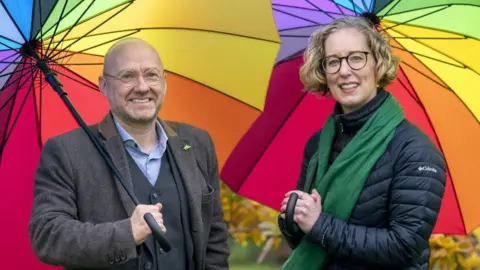 PA Media A bald man with glasses and a woman with blonde hair and glasses smile at the camera while standing under rainbow-coloured umbrellas. The man is wearing a grey suit and blue shirt, while the woman wears a dark jacket and green scarf 