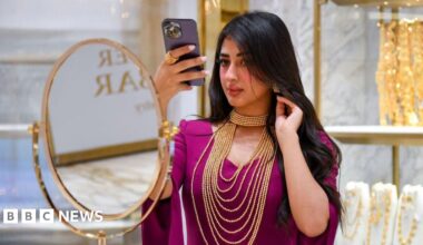 A woman checks herself out with a mobile phone as she tries a gold necklace during the 6th Edition of ASJAD Jewellery Exhibition at Alhazm Mall in Doha, Qatar, on 24 October 2024. She is wearing a purple sari traditional dress with gold earrings and gold chains around her neck.