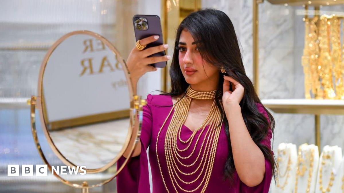 A woman checks herself out with a mobile phone as she tries a gold necklace during the 6th Edition of ASJAD Jewellery Exhibition at Alhazm Mall in Doha, Qatar, on 24 October 2024. She is wearing a purple sari traditional dress with gold earrings and gold chains around her neck.