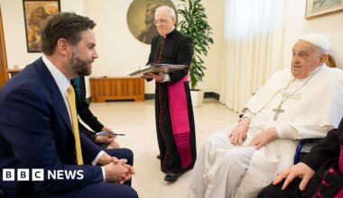 JD Vance wears a suit and sits in a chair in a beige room. He speaks to Pope Francis, who sits opposite him and wears white robes with a silver crucifix on a long chain