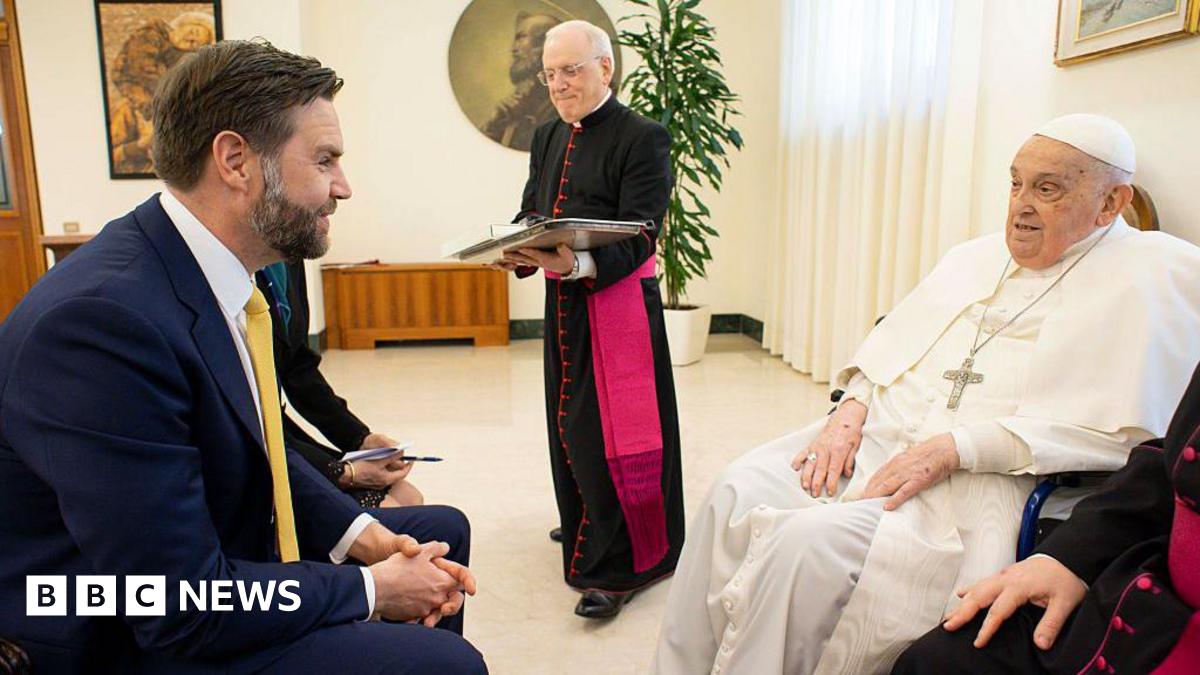 JD Vance wears a suit and sits in a chair in a beige room. He speaks to Pope Francis, who sits opposite him and wears white robes with a silver crucifix on a long chain