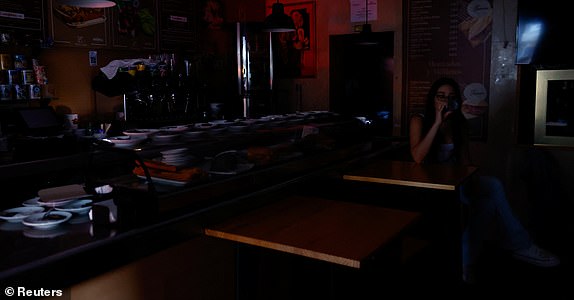 A woman has a drink in the dark inside a bar with outside light coming in through the door during a nationwide power outage in Madrid, Spain, April 28, 2025. The red glow is from a traffic light that recently came back on. REUTERS/Susana Vera