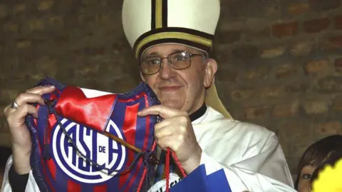 Getty Images Pope Francis is shown while a bishop in Buenos Aires. He is holding up a football pendant of his favourite team.