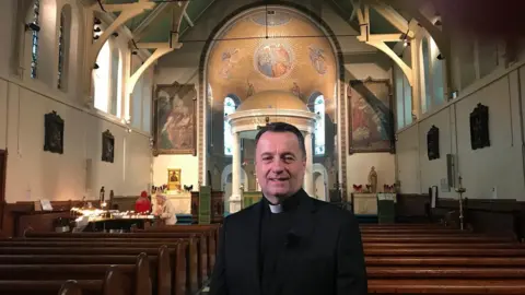 A man in traditional priest-wear smiles at the camera. He has short dark hair, and is standing in a church. Behind him is an alter, as well as paintings and other artwork on the wall.