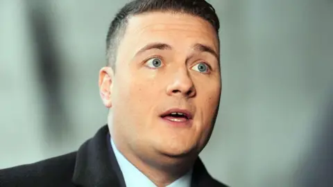 PA Media The health secretary Wes Streeting, a clean-shaven man with short dark hair, wears a light blue shirt and black coat, is speaking while looking away from the camera.