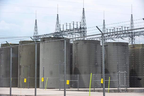 Fenced area with large cylindrical storage tanks and metal towers in the background, under a cloudy sky.