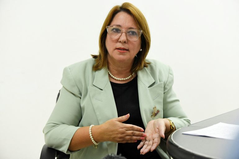 Maricela Sosa speaks in an interview in a white room. She wears a green blazer and glasses.