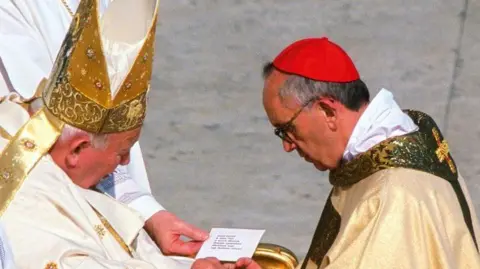 Getty Images Pope John Paul II and the future Pope Francis. Pope John Paul II is seated and his holding a card with writing on it. The future Pope Francis is dressed as a Cardinal and is kneeling before him.