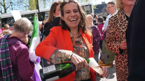 PA Media A smiling lady wearing a floral blouse and an orange blazer pours a glass of champagne on the street as people mill around in the background.