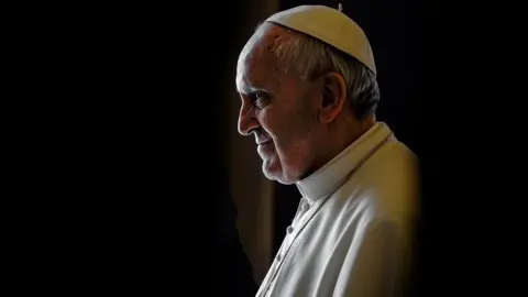 AP Pope Francis is shown in a head a shoulders shot.  He is wearing a white coat and white cap. He is shown in profile looking off the the left.