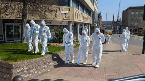 Alex Moss/BBC Group of people in white suits examining a patch of grass and a pavement