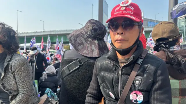 Lee, wearing a Yoon badge on his chest, looks at the camera through his sunglasses