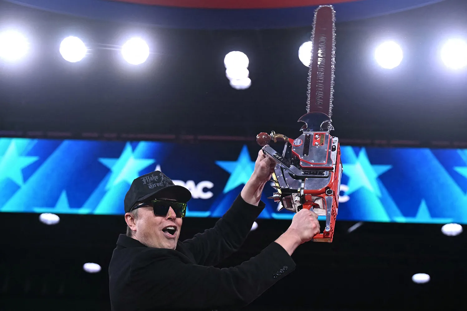 A man in sunglasses and a black MAGA cap yells as he holds up a chainsaw.