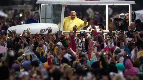 Getty  Pope Francis is seen wearing a yellow plastic rainproof in an open car. He is waving to the crowd which surrounds him.
