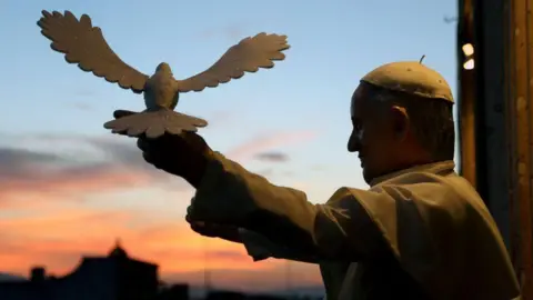 Getty Images Pope Francis poses with a model of a dove of peace. He holds his arm out and the model sits on his hand.