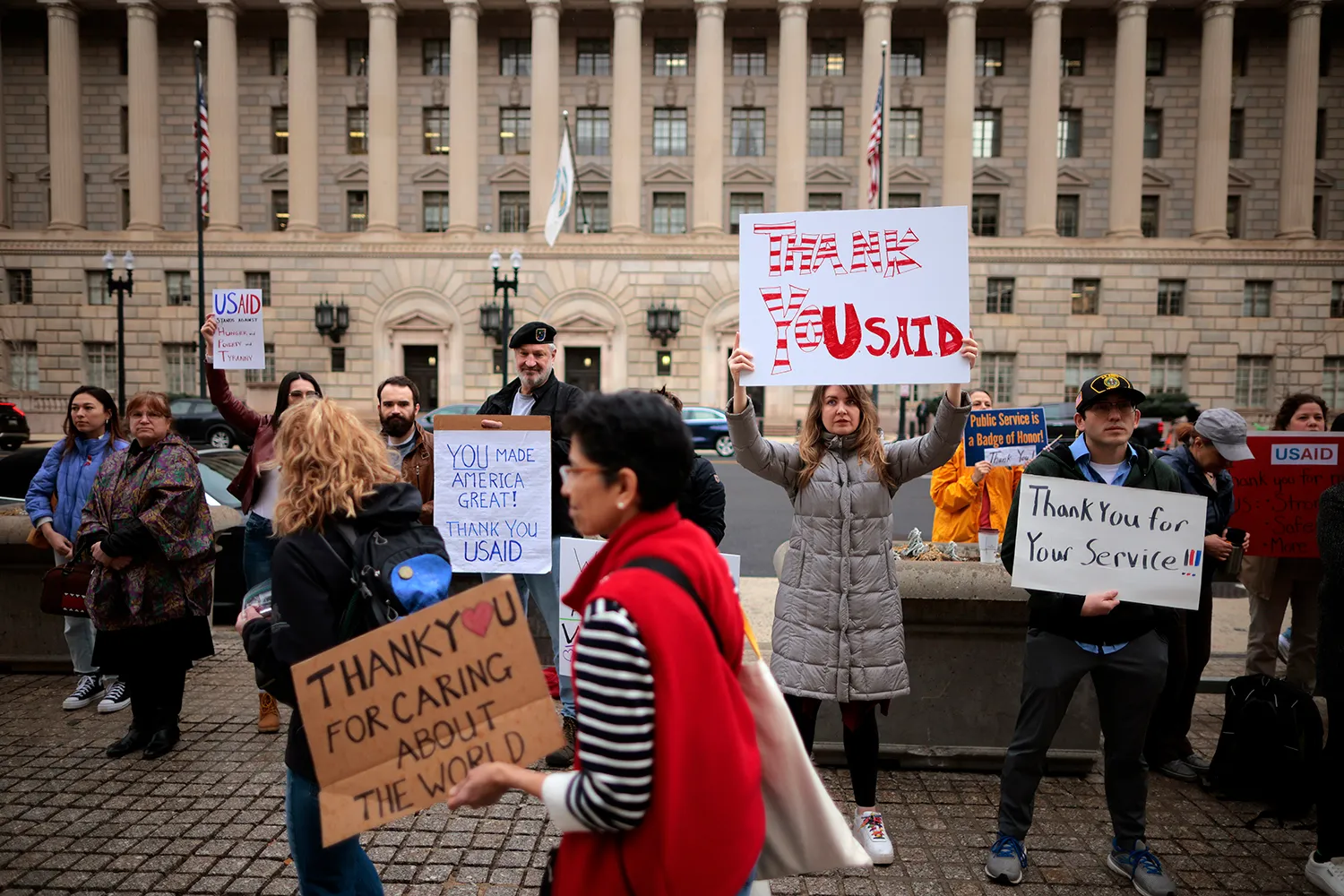People hold signs that say Thank You USAID and Thank you for caring for the world among other messages outside a federal building with columns.