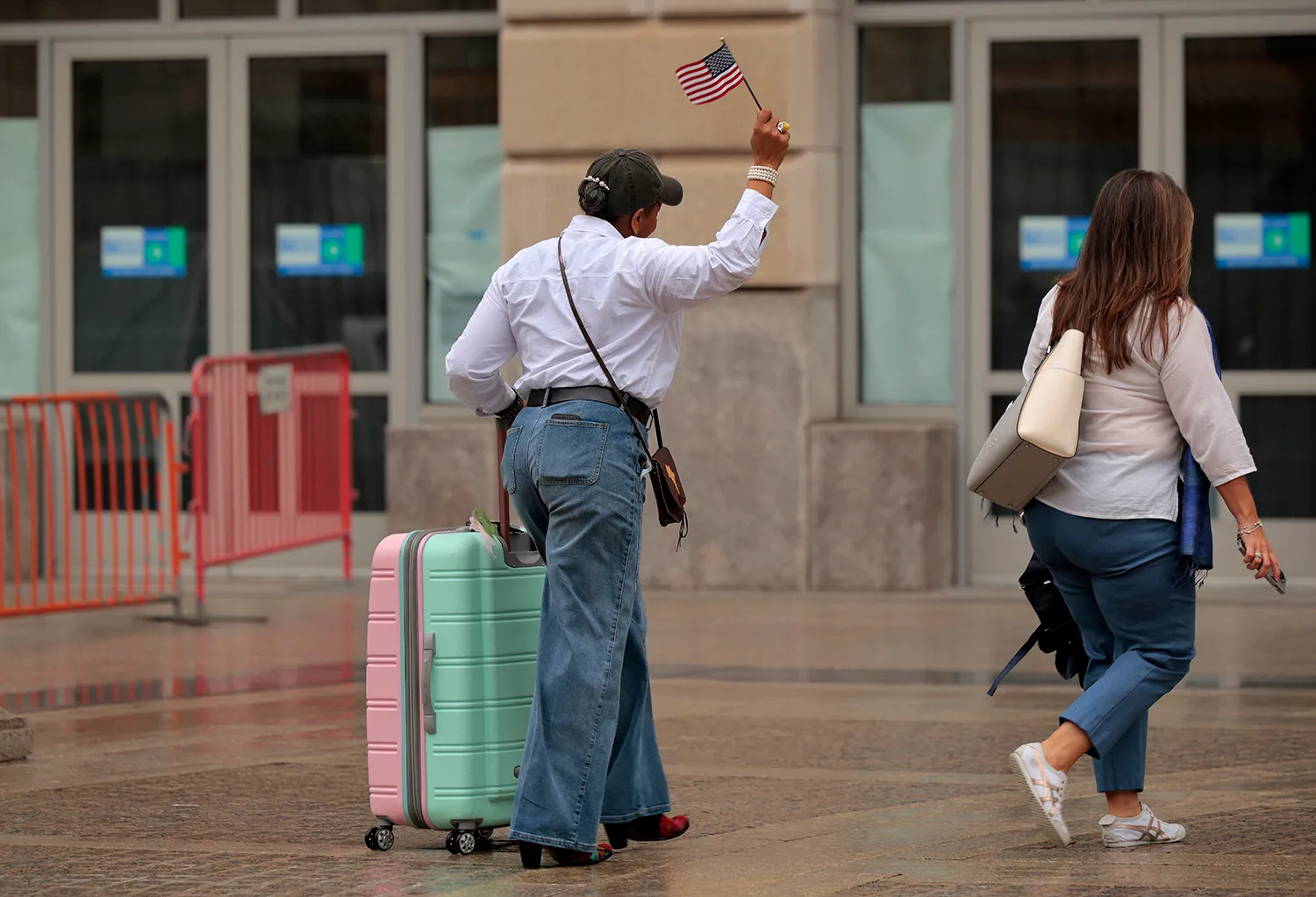 A person pulls a rolling bag and holds up a small U.S. flag as they walk away. Anotehr person walks ahead of her.