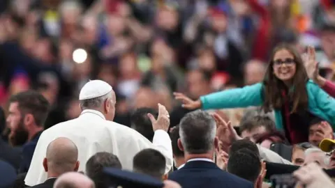 Getty Images A man wearing white papal gowns and a white Zucchetto (small hat) is amongst a crowd of people who are reaching out towards him.