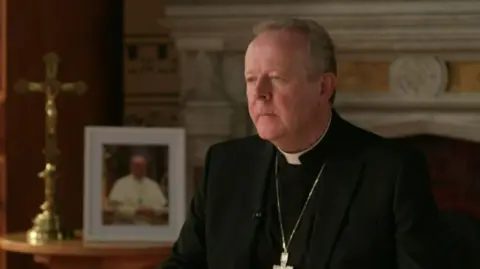 A man wearing traditional Catholic priest-wear sits, looking to the left of the camera. Behind him, there is a framed photograph of the Pope.