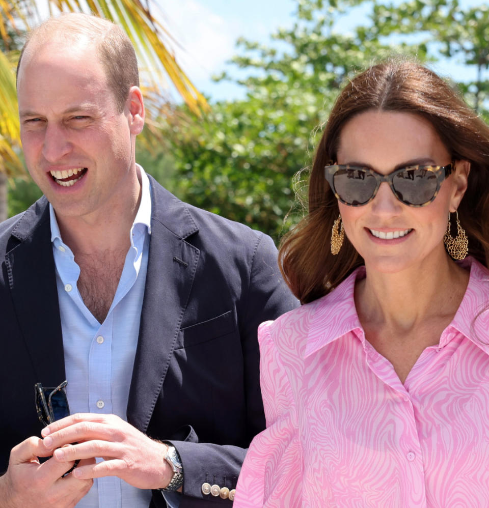 Prince William wearing a blue open necked shirt with a navy blazer standing outside next to Kate Middleton in a pink dress and gold hoop earrings with sunglasses.