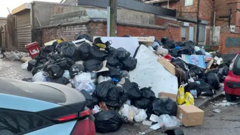 A large pile of rubbish, including black bin bags, boxes and a mattress, on a pavement. The pile is spilling out onto the road.