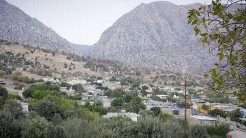 Phil Caller A panoramic shot of an attractive village with small houses and other buildings nestled among trees, greenery and farming land. Mountains loom over the village 