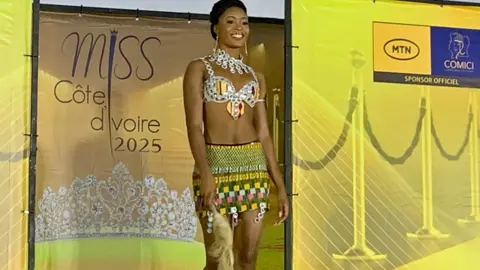 A beauty pageant contestant in Daloa wearing a bejewelled bikini and short shirt with traditional designs and holding a fly-whisk walks on to the catwalk. Behind her are banners in yellow with the Miss Cote d'Ivoire 2025 logo. 