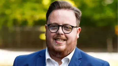 Getty Images Thomas Kerr, a man with dark glasses, light brown hair and a ginger beard, smiles in a close-up shot. He is wearing a blue suit and white shirt, with blurred-out vegetation in the background