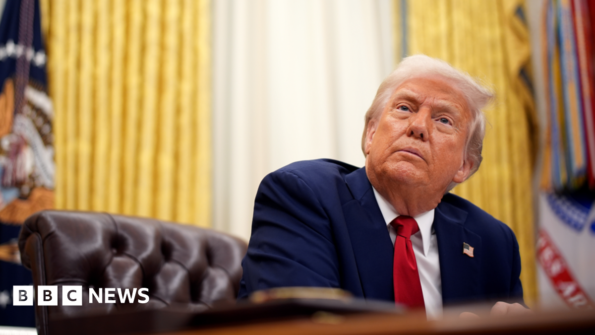 Donald Trump in the Oval Office sitting at his desk, looking up. He's wearing a navy blue suit, red tie, has an US flag pin on his jacket, and there are big gold curtains hanging behind him