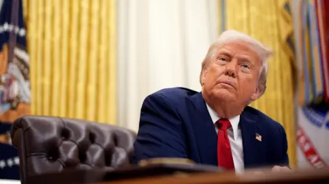 Getty Images Donald Trump in the Oval Office sitting at his desk, looking up. He's wearing a navy blue suit, red tie, has an US flag pin on his jacket, and there are big gold curtains hanging behind him