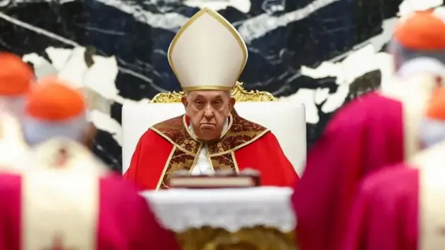 Pope Francis in red robes as he sits while cardinals look at him