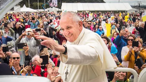 PA A man in the foreground of the picture, dressed in white papal vestments with a large cross on a chain around his neck, waves to a huge crowd of people, who can be seen in the background. Many are holding aloft cameras. 