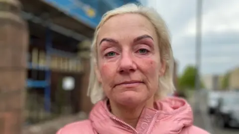 A woman with blonde hair looks at the camera. Her eyes are red maybe because she has been crying. She is wearing a pink jacket and standing on a street, with a blurred building, cars and a lamppost behind her. 